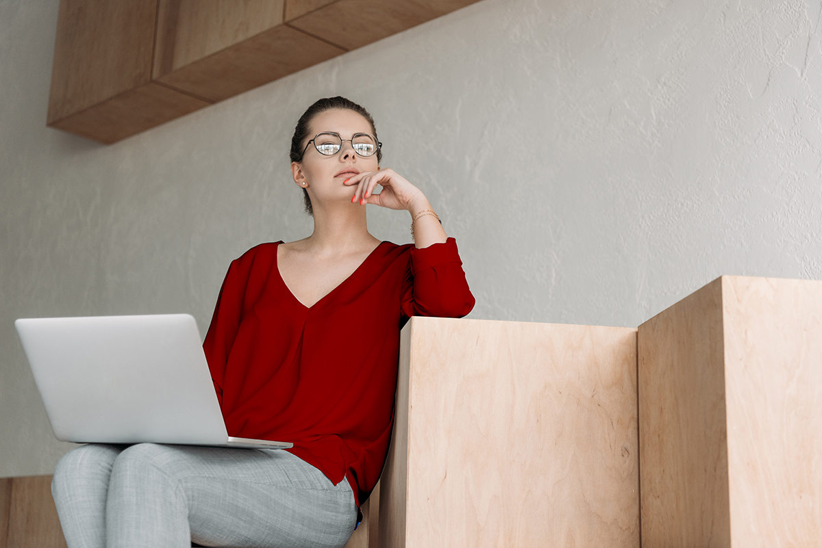 Attractive thoughtful fashionable businesswoman working with laptop while sitting on wooden cubes
